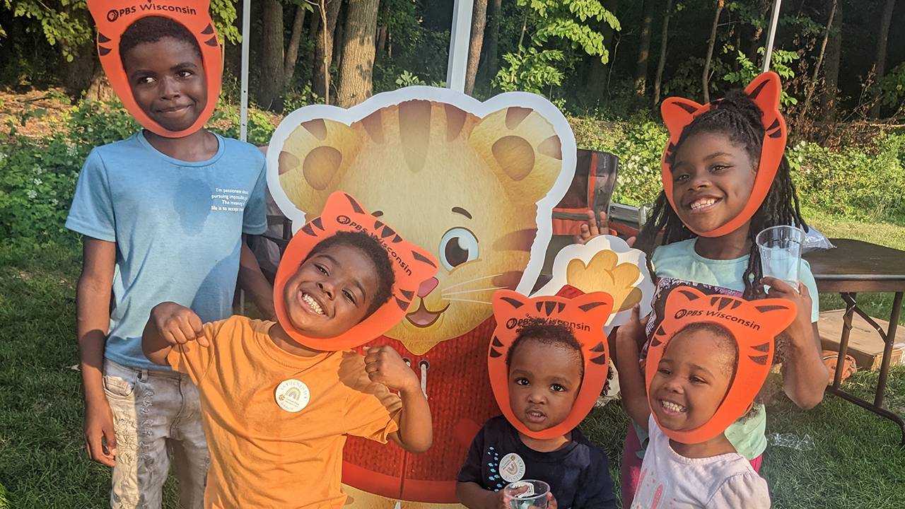 Six smiling children wearing orange PBS Wisconsin tiger ear headbands pose outdoors with a large Daniel Tiger cutout.