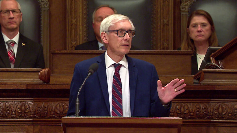 White-haired man in a navy suit and striped tie speaks at a podium in a formal wood-paneled government chamber, with officials seated behind him.