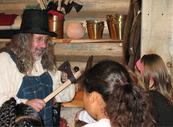 A man in fur trade era clothes talks to children on a historical tour at a museum.