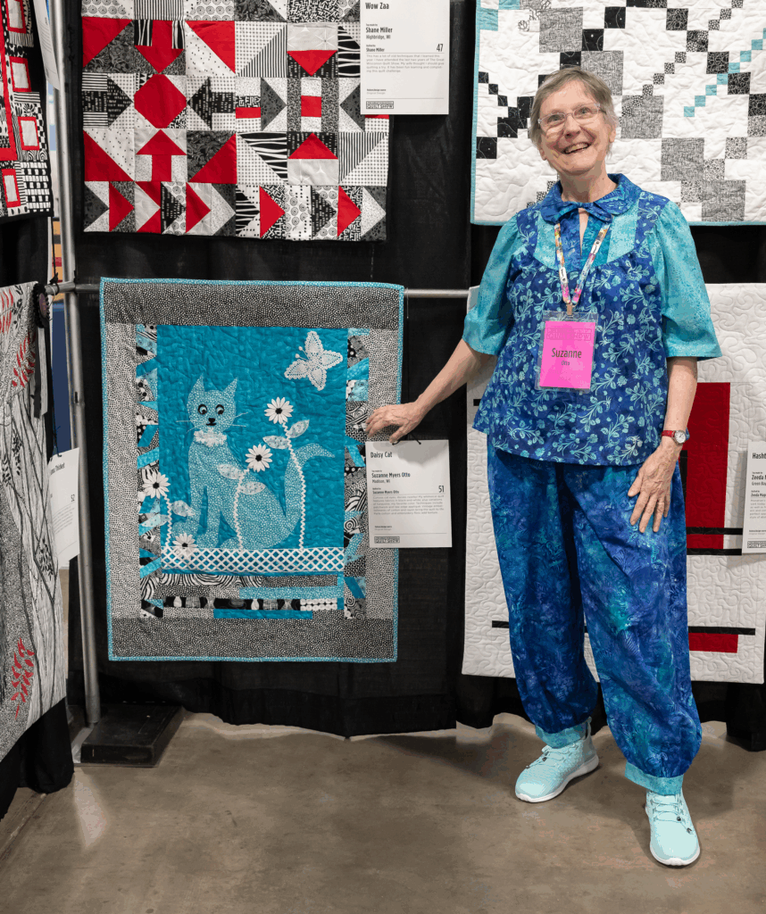 A woman in blue stands beside her turquoise and black quilt featuring a stylized cat, flower, and butterfly design.