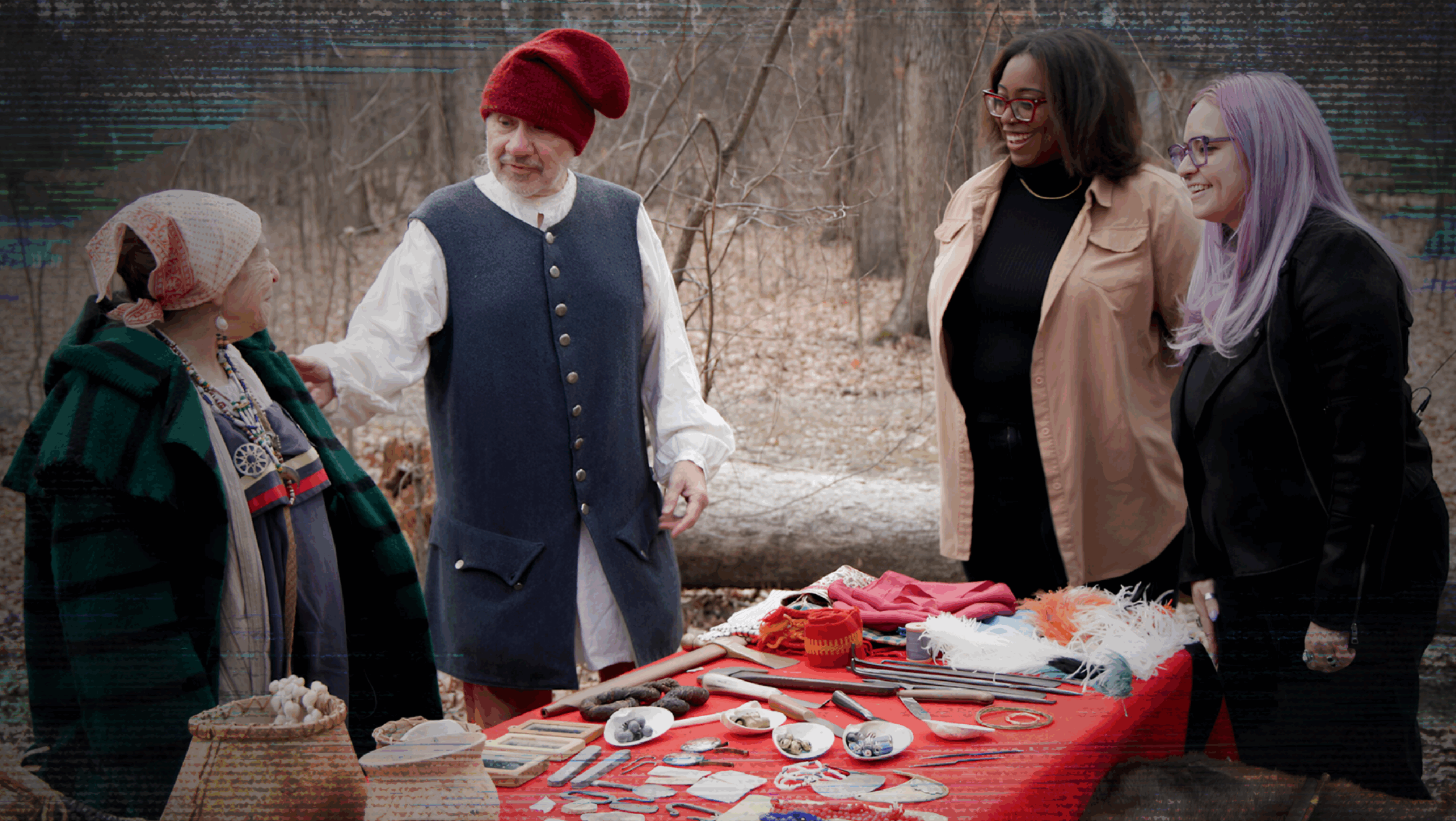Four people stand around a table outside. The woman and man on the left are dressed in fur trade era clothing. The two women on the right are wearing modern-day clothes.