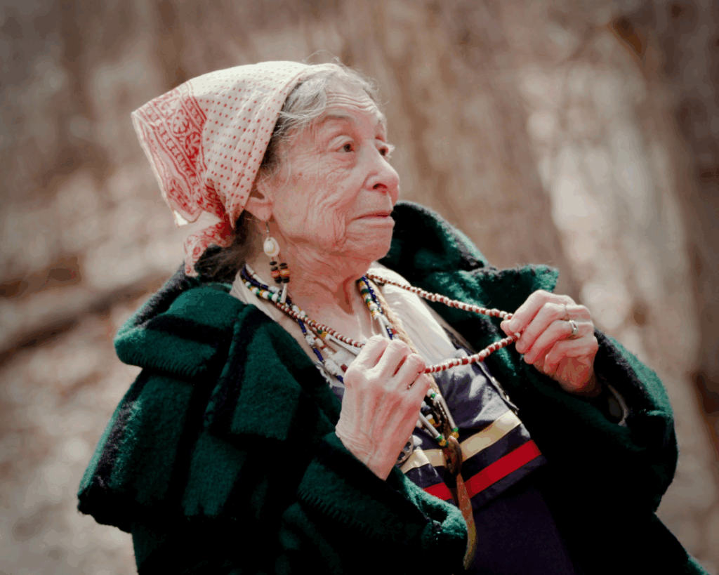 A woman in fur trade era clothing holds up a necklace she's wearing.