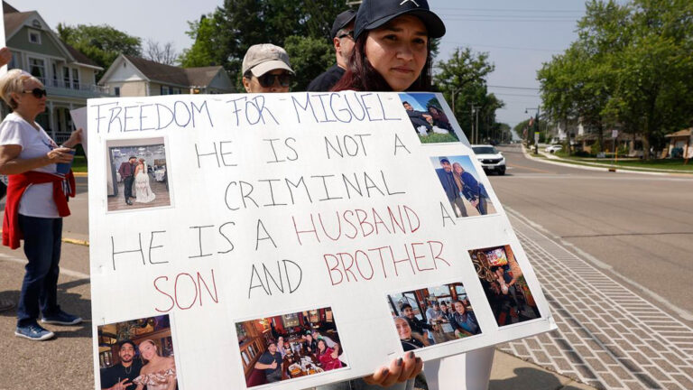 Young woman holding a protest sign reading “FREEDOM FOR MIGUEL. HE IS NOT A CRIMINAL. HE IS A HUSBAND, A SON AND BROTHER,” with family photos.