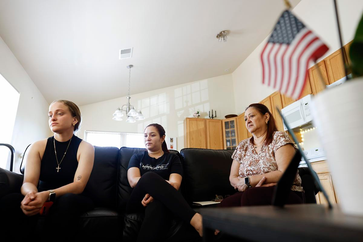 Three women seated on a black couch in a well-lit home, appearing somber, with a small American flag in the foreground.