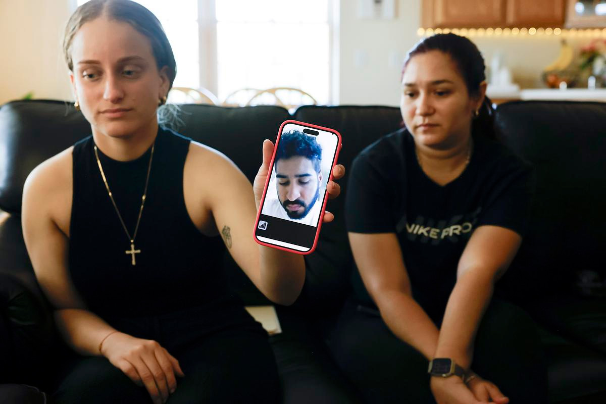 Young woman holding a phone showing a video call with a man, while seated beside another woman on a black couch.