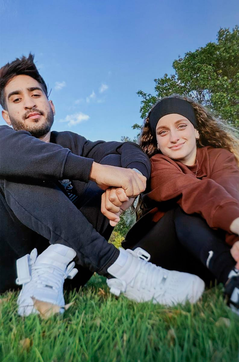 “Young couple sitting on grass, holding hands, both wearing dark clothes and white sneakers, with blue sky and trees behind them.”