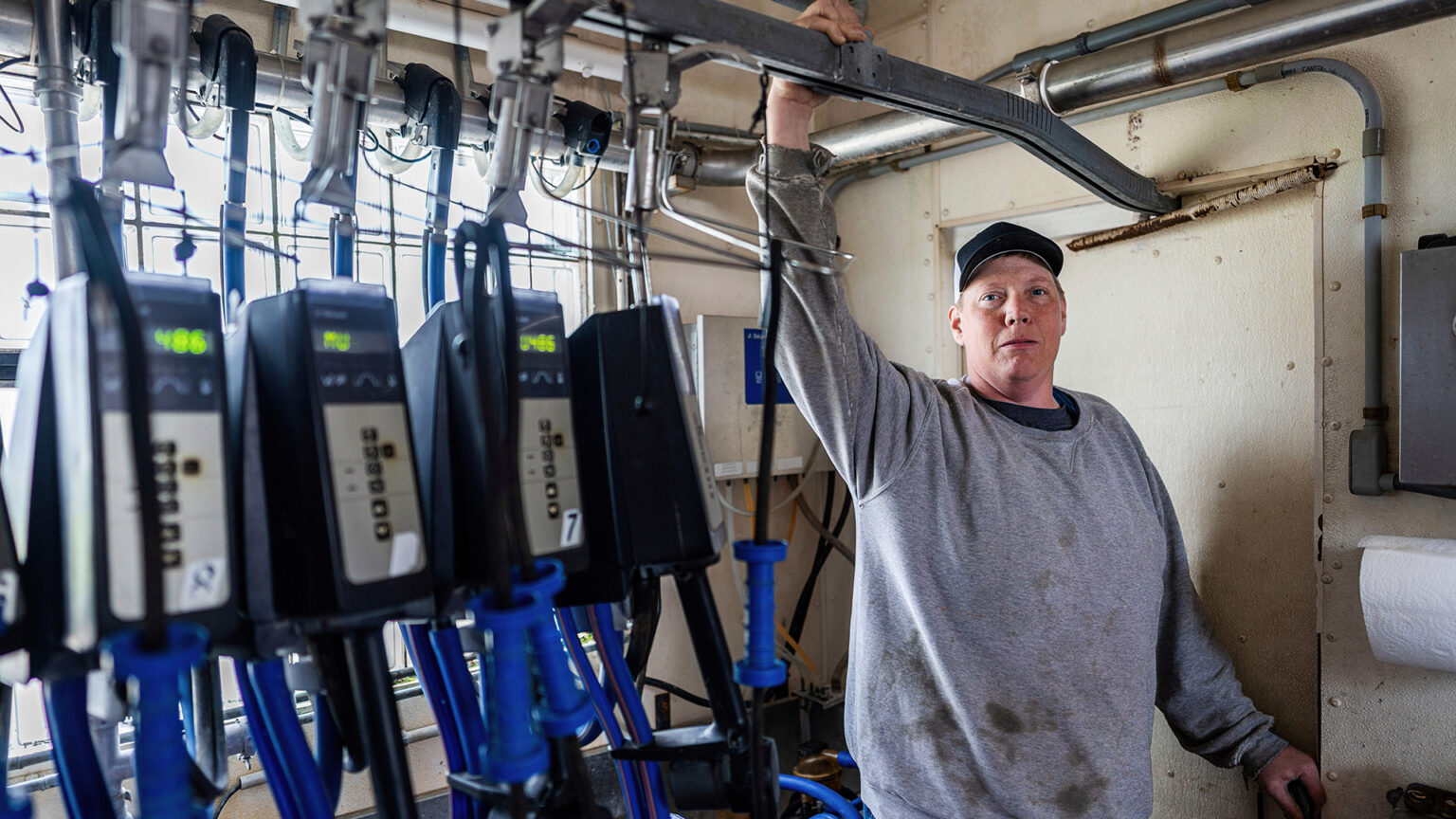 Adam Faust stands in a room with his right arm holding an overhead brace for a milking system and his left hand holding a door handle, with a daylight illuminated window in the background and multiple digital gauges in a row in the foreground. Adam Faust stands in a room with his right arm holding an overhead brace for a milking system and his left hand holding a door handle, with a daylight illuminated window in the background and multiple digital gauges in a row in the foreground.