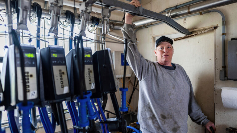 Adam Faust stands in a room with his right arm holding an overhead brace for a milking system and his left hand holding a door handle, with a daylight illuminated window in the background and multiple digital gauges in a row in the foreground.