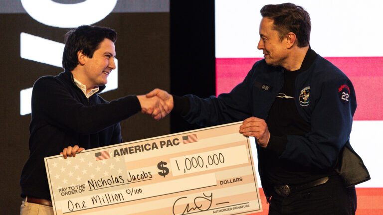 Nicholas Jacobs and Elon Musk stand and shake their right hands while their left hands hold an oversized paper check with the words America PAC, pay to the order of Nicholas Jacobs and $1,000,000, with an illuminated screen in the background.