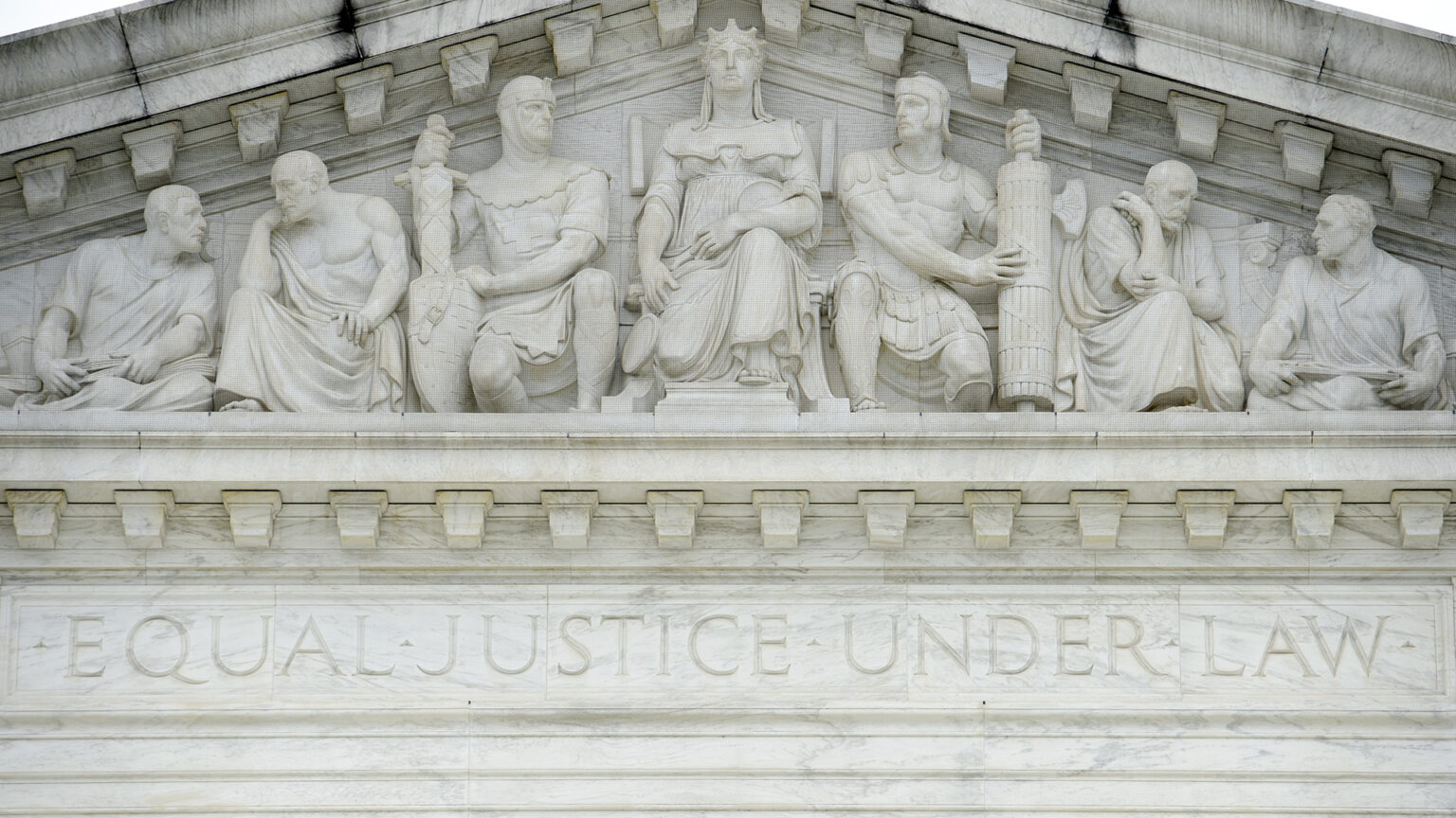 A pedimental frieze showing statues of seven figures seated above the words Equal Justice Under Law is carved in a marble masonry building. A pedimental frieze showing statues of seven figures seated above the words Equal Justice Under Law is carved in a marble masonry building.