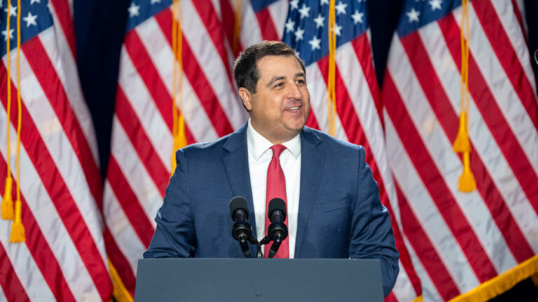 Man in blue suit and red tie speaking at a podium with microphones, standing before a backdrop of multiple U.S. flags.