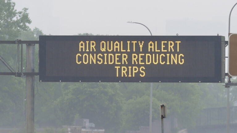 An electronic traffic message sign mounted on metal poles over a roadway reads Air Quality Alert Consider Reducing Trips, with trees, light poles and buildings in the background obscured by haze.