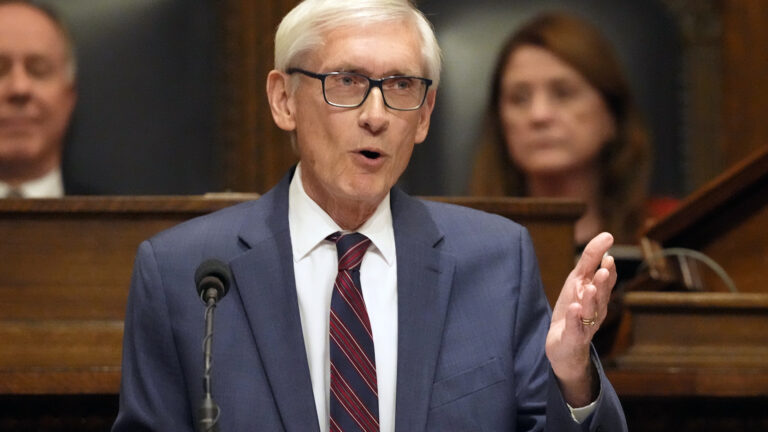 Tony Evers gestures with his left hand while standing and speaking into a microphone, with two out-of-focus people seated in high-backed leather and wood chairs on a wood legislative dais in the background.