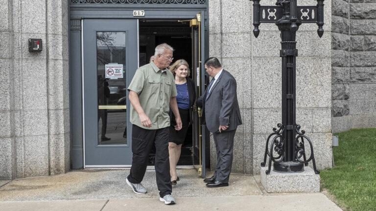 Hannah Dugan walks away from an open door, with another person walking in front of her and another person holding the door open for her, in front of a stone masonry building and next to a metal light fixture.