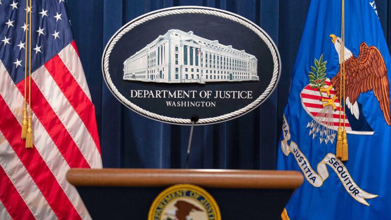 An oval logo showing an illustration of a multi-story Beaux Arts-style building with a U.S. flag on a flagpole in front of a colonnade is mounted in front of a stage curtain and between a U.S. flag and the U.S. Department of Justice flag, with an  out-of-focus podium with a microphone mounted to its surface in the foreground.