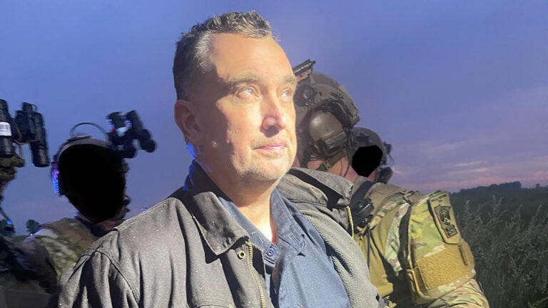 Vance Luther Boelter stands outdoors with a spotlight illuminating his face, with multiple law enforcement officers wearing camouflage uniforms and helmets with headphones and mounted scopes standing in the background with black areas redacting their faces, with foliage and a tree line on the horizon under a cloudy sky at dusk.