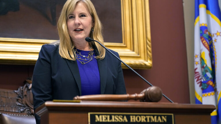 Melissa Hortman stands and speaks into a microphone mounted to the top of a wood podium with an out-of-focus wood gavel on its surface and a name plate reading Melissa Hortman on its front, with a wood-carved chair, framed painting and Minnesota flag in the background.