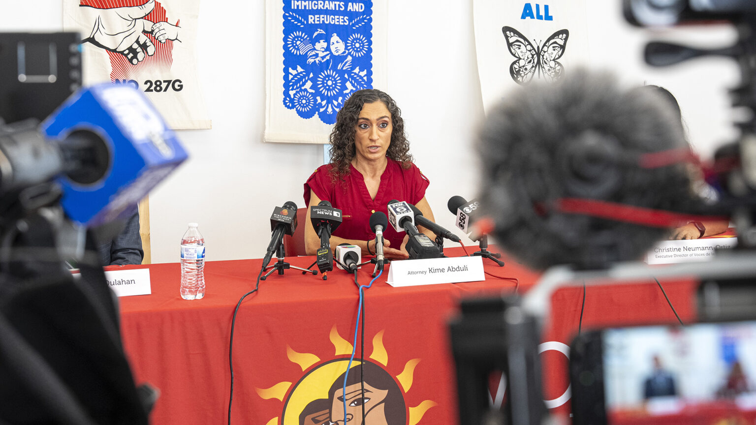 Kime Abduli sits at a table and speak into multiple microphones with the flags of different media organizations mounted on short tripods set on its surface along with a water bottle and paper name tents in a room with prints displayed on a wall in the background, with out-of-focus video camera microphones and monitors in the foreground. Kime Abduli sits at a table and speak into multiple microphones with the flags of different media organizations mounted on short tripods set on its surface along with a water bottle and paper name tents in a room with prints displayed on a wall in the background, with out-of-focus video camera microphones and monitors in the foreground.