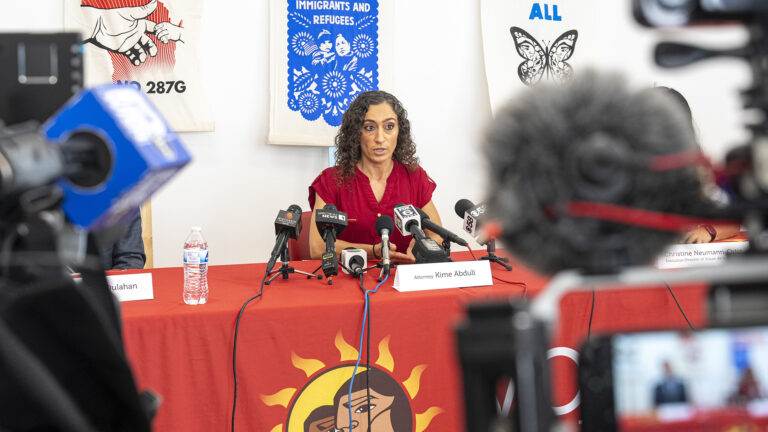 Kime Abduli sits at a table and speak into multiple microphones with the flags of different media organizations mounted on short tripods set on its surface along with a water bottle and paper name tents in a room with prints displayed on a wall in the background, with out-of-focus video camera microphones and monitors in the foreground.