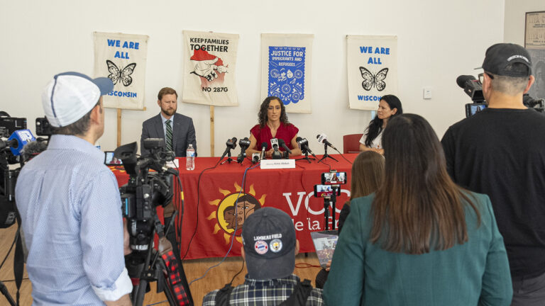 Cain Oulahan, Kime Abduli and Christine Neumann-Ortiz sit at a table and speak into multiple microphones with the flags of different media organizations mounted on short tripods set on its surface along with a water bottle and paper name tents in a room with prints displayed on a wall in the background, with seated and standing people in the foreground with video cameras and cell phones mounted on tripods facing them in the foreground.