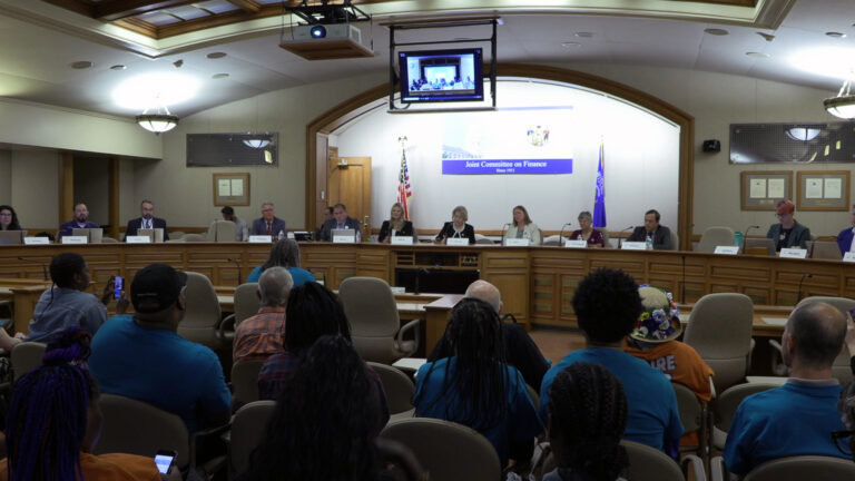 Multiple sit behind mounted microphones and paper printed name plates on top of a wood-paneled legislative dais with a TV monitor mounted overhead under a vaulted ceiling, with U.S. and Wisconsin flags in the background, with people seated in multiple rows of stacking chairs facing the dais in the foreground.