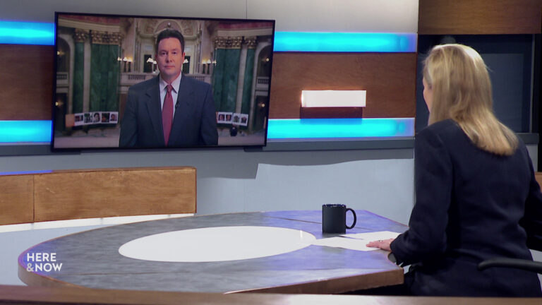 Frederica Freyberg sits at a desk on the Here & Now set and faces a video monitor showing an image of Zac Schultz.