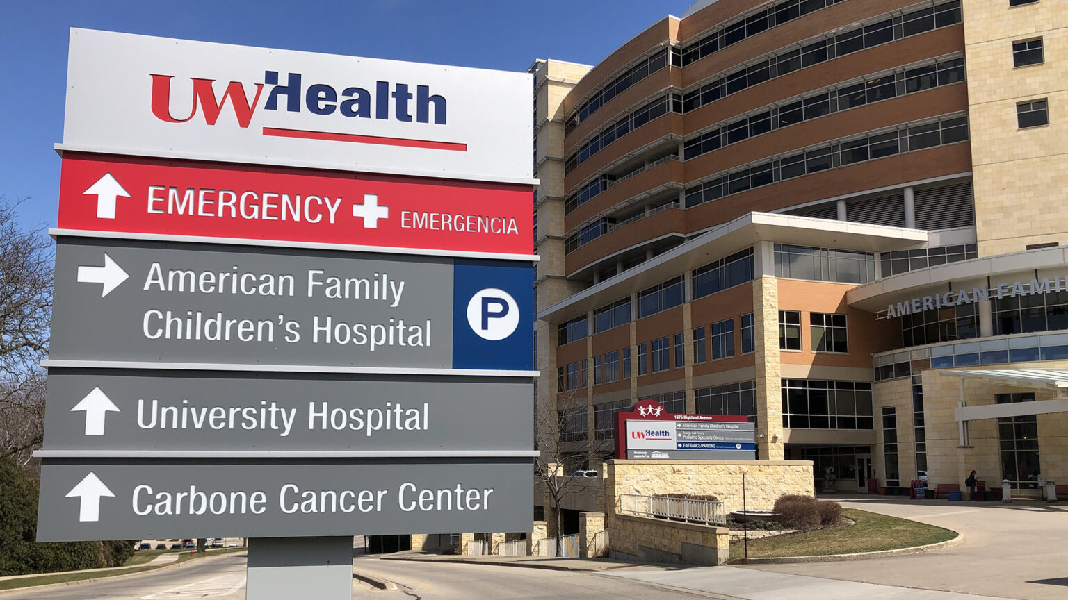 A multi-tier sign with the UW Health wordmark, the words Emergency + Emergencia, American Family Children's Hospital and P, University Hospital and Carbone Cancer Center with arrows pointing in different direction stands in a median of a road next to a driveway to a multistory building with a stone masonry and brick exterior, rows of windows of different sizes and more signs. A multi-tier sign with the UW Health wordmark, the words Emergency + Emergencia, American Family Children's Hospital and P, University Hospital and Carbone Cancer Center with arrows pointing in different direction stands in a median of a road next to a driveway to a multistory building with a stone masonry and brick exterior, rows of windows of different sizes and more signs.