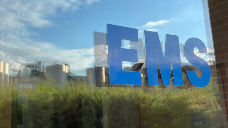 Blue “EMS” letters on a glass window reflecting a partly cloudy sky, green trees, and industrial buildings in the background.