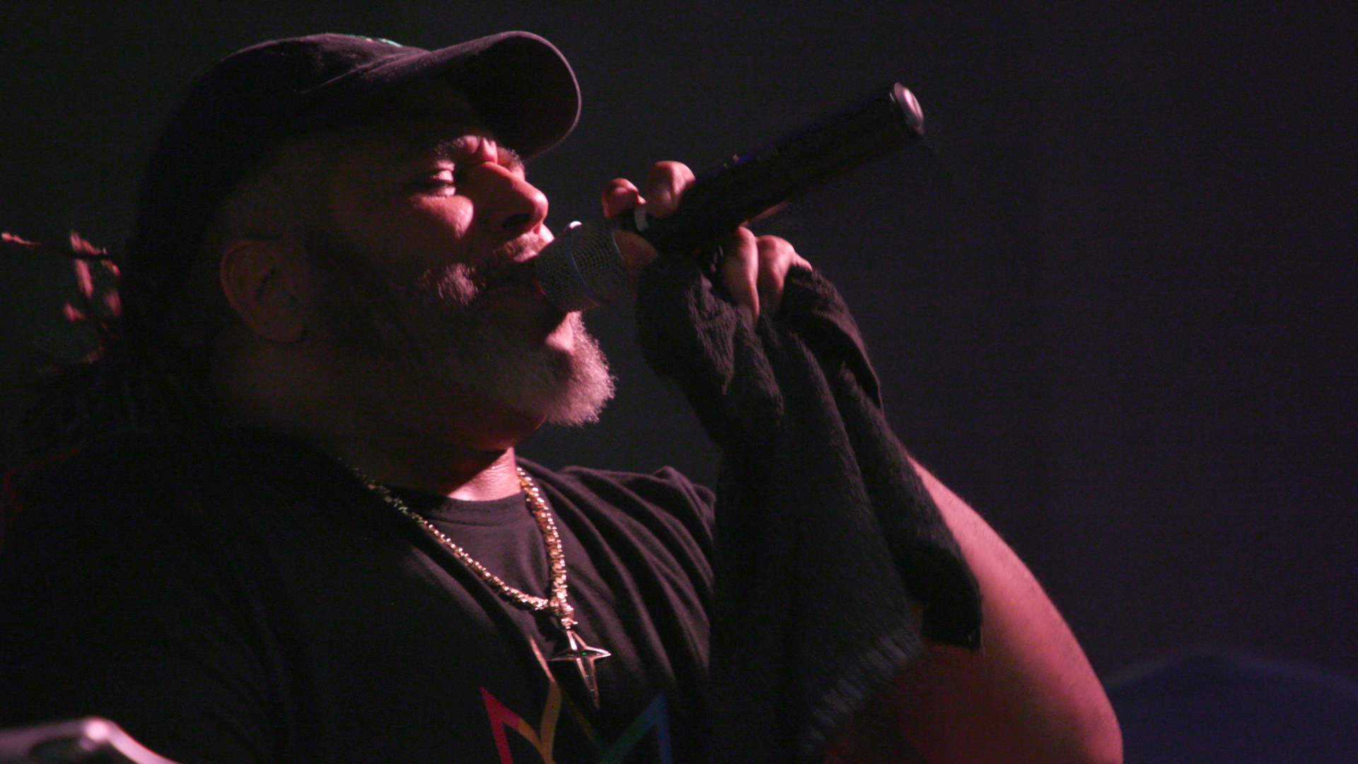 A musician performs passionately on stage, holding a microphone close to his mouth. He wears a black cap, a black shirt and chain necklace. The “Mile of Music” logo is displayed in the lower right corner.