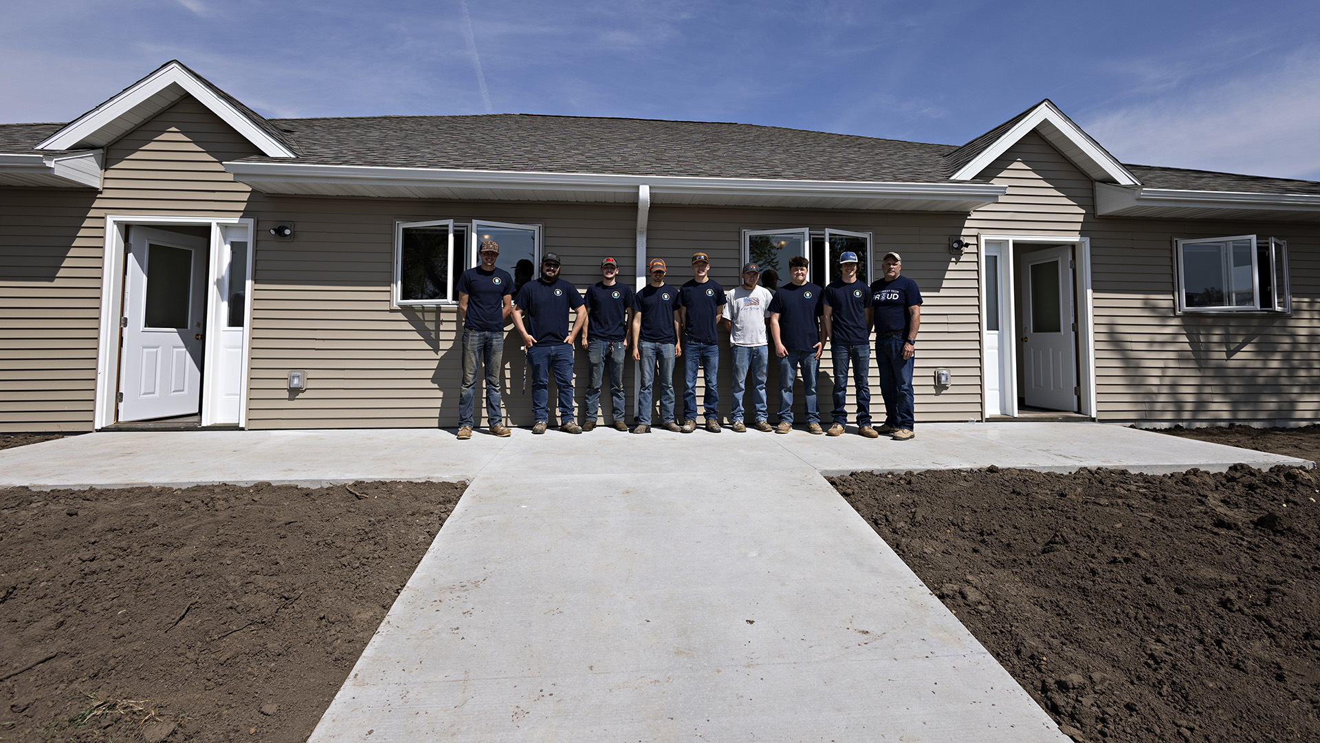 Nine students stand on a concrete patio in front of a single-story duplex with a mirror design of doors and windows, with bare dirt and a sidewalk in the foreground.