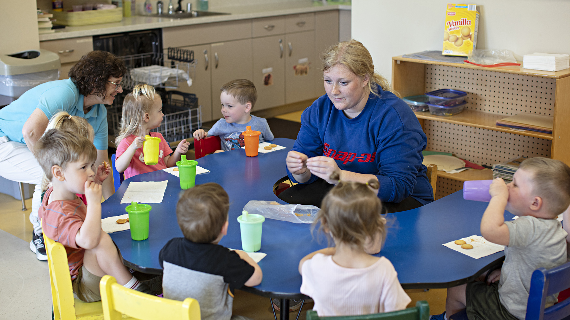 Grace Kite sits at the center of a low table in the shape of a half-circle, with seven young children siting in low chairs around its outer circumference while eating and drinking snacks, with Paula Timmerman siting in a chair to the side, in a room with shelves, cabinets and an open dishwasher in the background.