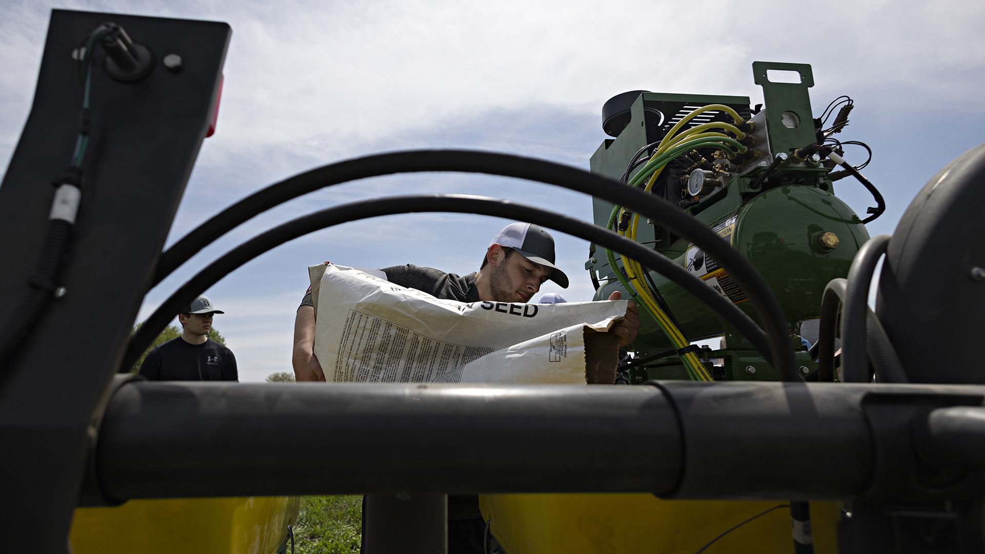 A student holds up a large paper bag with one open end to fill a container in a farm vehicle, with another student standing in the background.