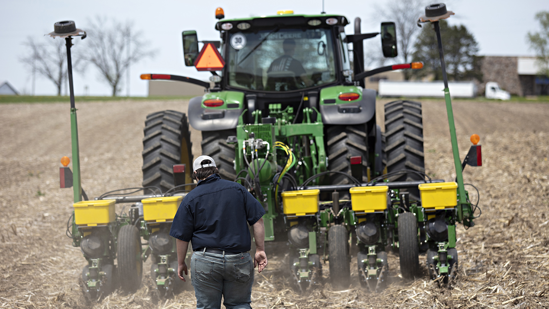 Parker Reese walks in the dirt and straw of a field behind a tractor pulling a planter, with out-of-focus buildings and a parked semi-truck on the horizon in the background.