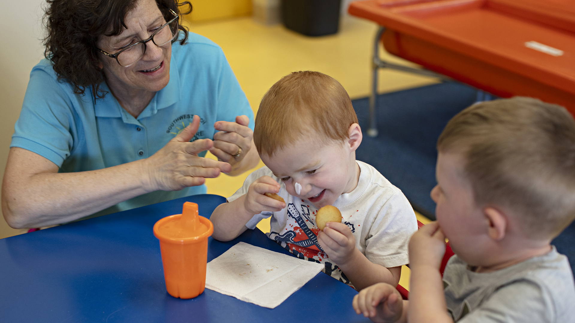 Paula Timmerman dabs sunscreen on her fingers while crouching next to a low table where two children are seated, one with sunscreen on their nose, with a sippy cup and napkin on its surface.