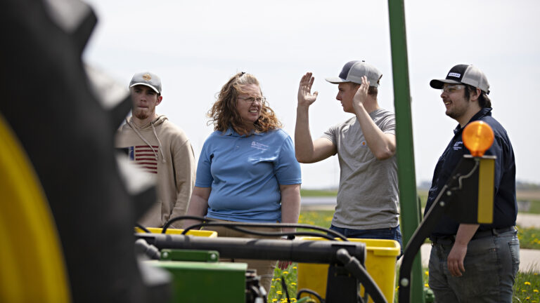 Christina Winch stands outdoors and speaks to Jamin Crapp, who holds both hands up in the air, with two other people standing on either side of them, with an out-of-focus wheel and chassis of a farm vehicle in the foreground.