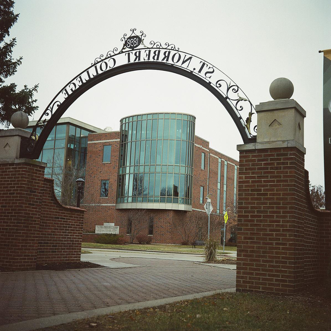 A filigree metal arch with the words "St. Norbert College" as seen from the rear connects two brick walls topped by concrete pillars with spherical tops over a brick walkway, with a multistory brick and glass-walled building in the background.