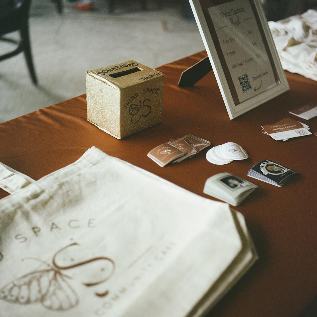 Canvas tote bags, stickers and t-shirts are stacked on a table with a tablecloth along with a framed sign showing prices and cardboard box with an open slot in its top and the words "Donations," "Thank You" and "Third Space" written on its surface.