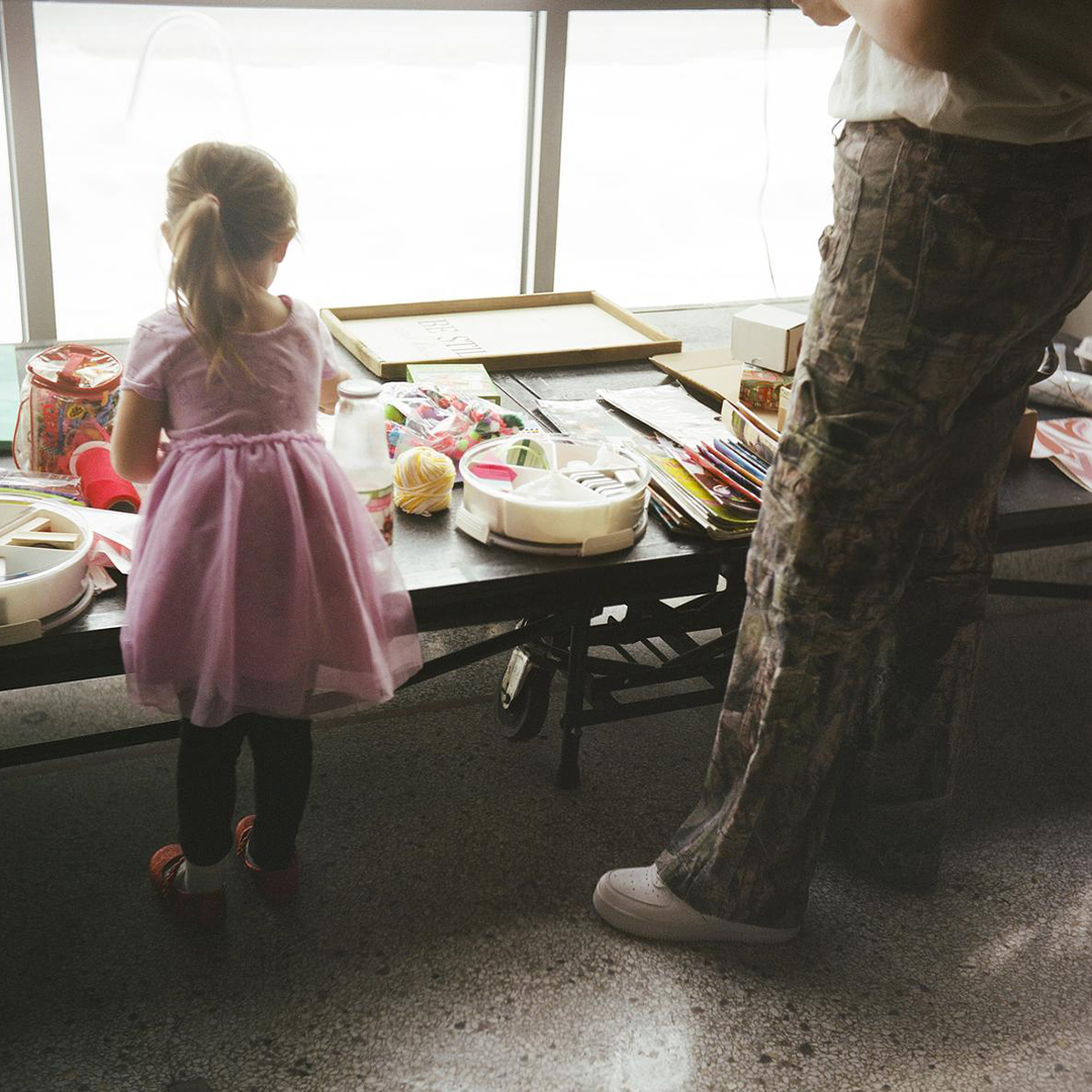 A child and Jasmine Gordon lean over a low table with multiple items on its surface while standing on a polished concrete floor illuminated by plate-glass windows.