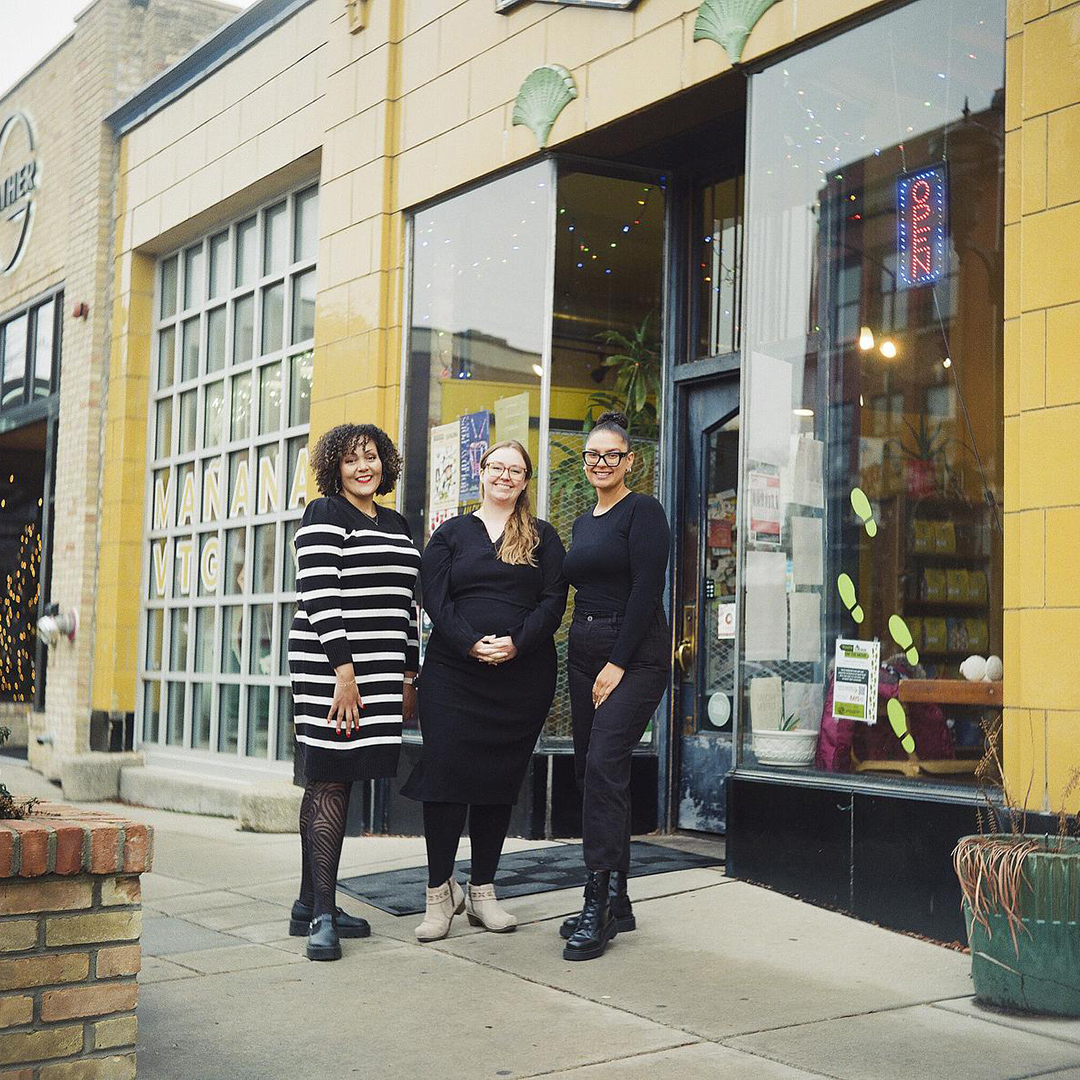 Ivy McGee, Sarah Titus and Jasmine Gordon pose for a portrait while standing on a sidewalk outside the entrance of a building with large plate-glass windows and an illuminated "Open" sign.