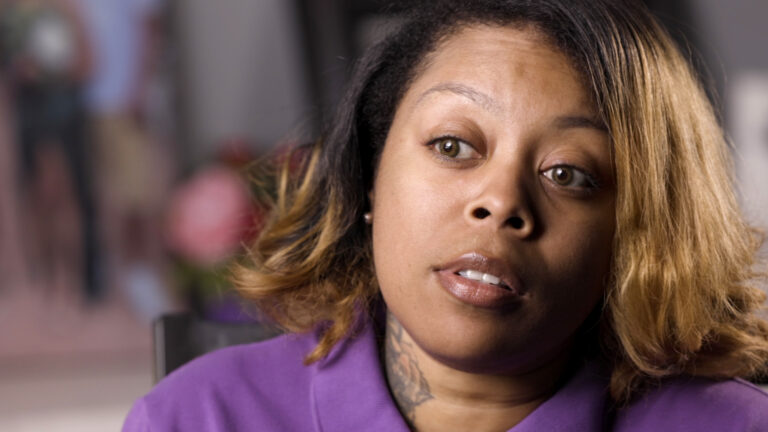 A Black woman with shoulder-length hair and a neck tattoo wearing a purple shirt, looking thoughtfully past the camera.