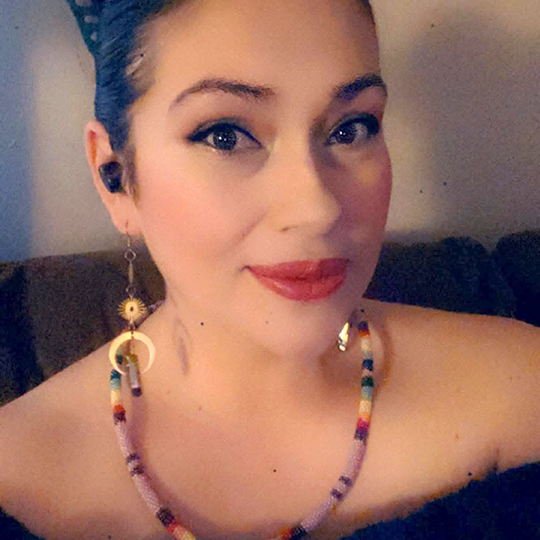 Deidre Ann Caldwell - Beading Advisor - Powwow Bound Woman with dark hair in a bun, wearing a colorful beaded necklace and earrings, smiling in indoor lighting.