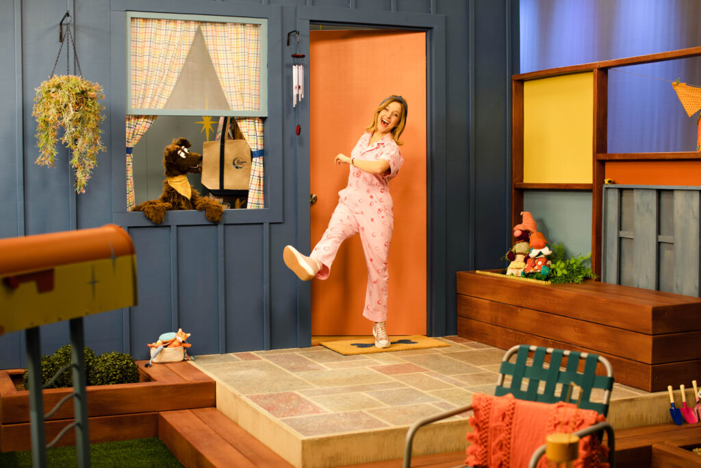 A woman in a pink jumpsuit steps outside of a blue house with a puppet dog in the window.