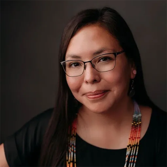 Jennifer Gauthier - Cultural Advisor - Powwow Bound Woman with long dark hair and glasses, wearing a black shirt and a multicolored beaded necklace, smiling softly against a dark background.