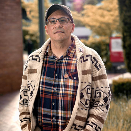 Justin Eagle Gauthier - Narrative Advisor - Powwow Bound Man wearing glasses, a cap, a plaid shirt, and a patterned cardigan, standing outside in an urban setting with trees in the background.