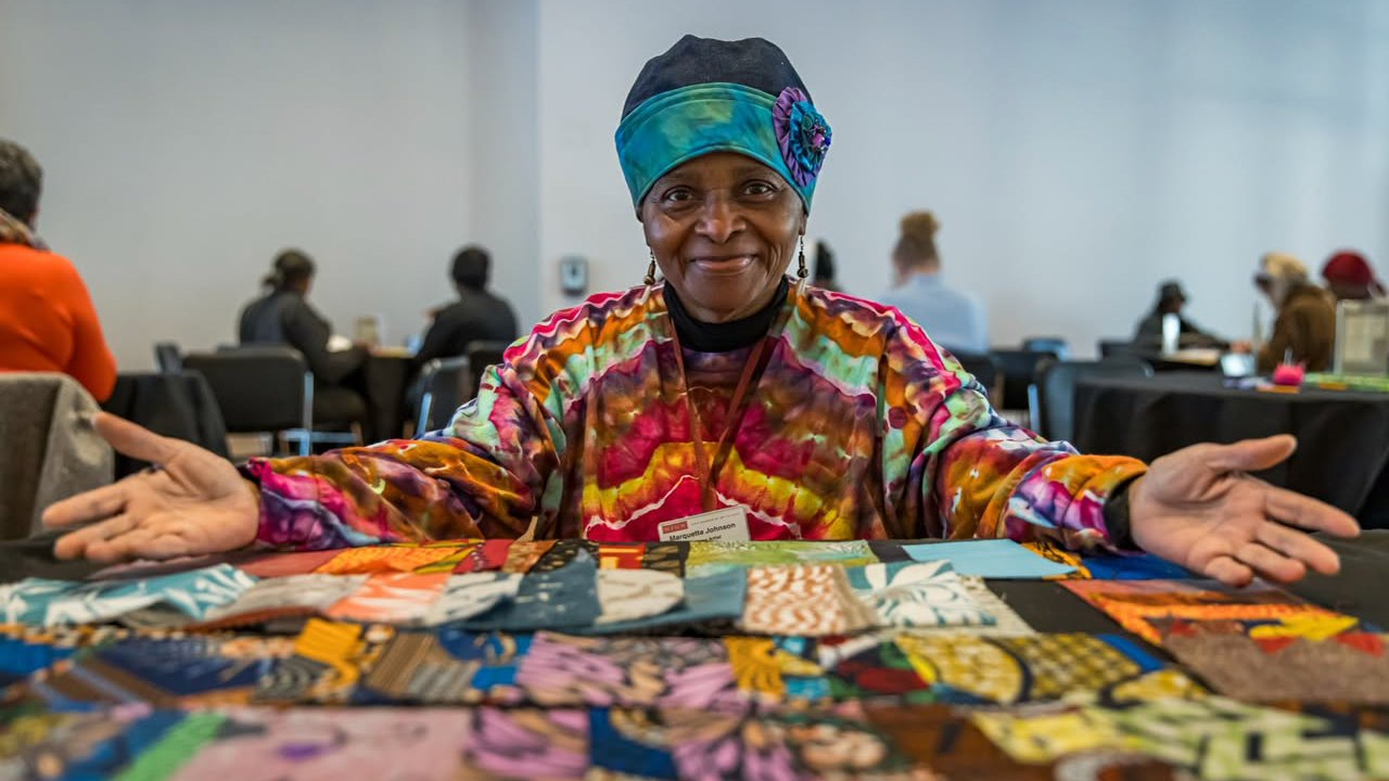 Smiling woman in colorful attire sits at a table displaying vibrant fabric squares, arms open wide, in a room with others working in the background.