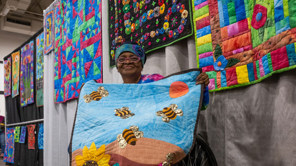 A woman in a wheelchair proudly holds up a quilt featuring sunflowers and bees, with vibrant quilts displayed behind her.