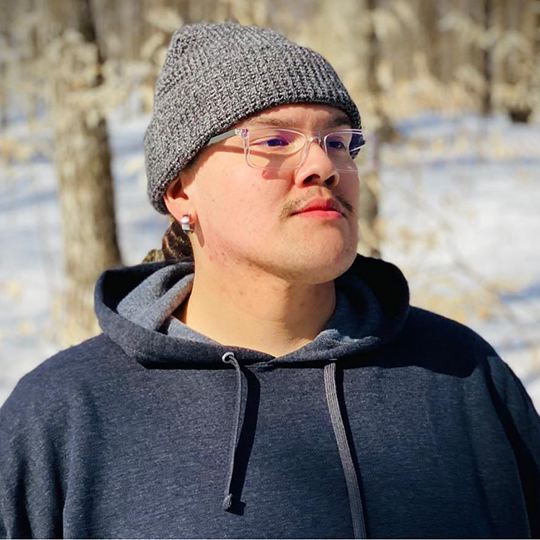 Myron Pyawasit - Traditional Drum Music - Powwow Bound Person with a mustache wearing a gray beanie and dark hoodie, standing in a snowy forest with trees in the background.