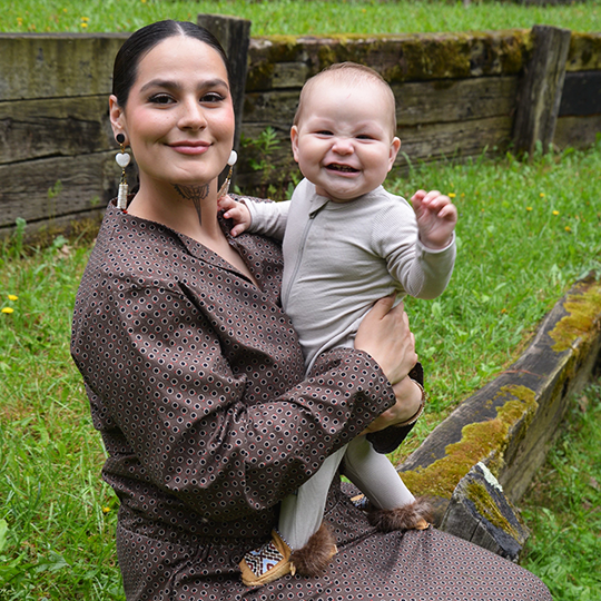 Naneque LaTender - Menominee Language Advisor - Powwow Bound Woman wearing a brown patterned dress and heart-shaped earrings, holding a smiling baby dressed in beige, seated outdoors on a wooden structure.