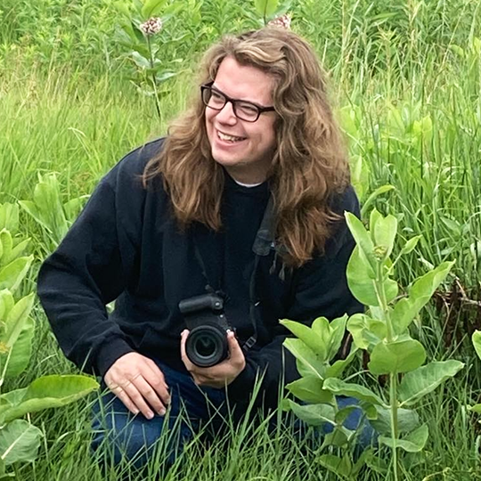 Nicholas Schwitzer - Character Concept Artist - Powwow Bound Person with long wavy hair and glasses, kneeling in a grassy field while holding a camera and smiling.