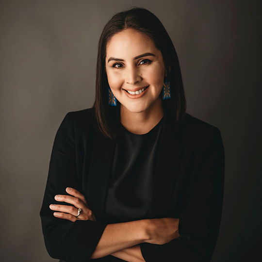 Pamela Boivin - Women's Fancy Shawl Dance Advisor - Powwow Bound Woman with shoulder-length dark hair, wearing a black outfit and blue beaded earrings, smiling with arms crossed against a dark studio background.